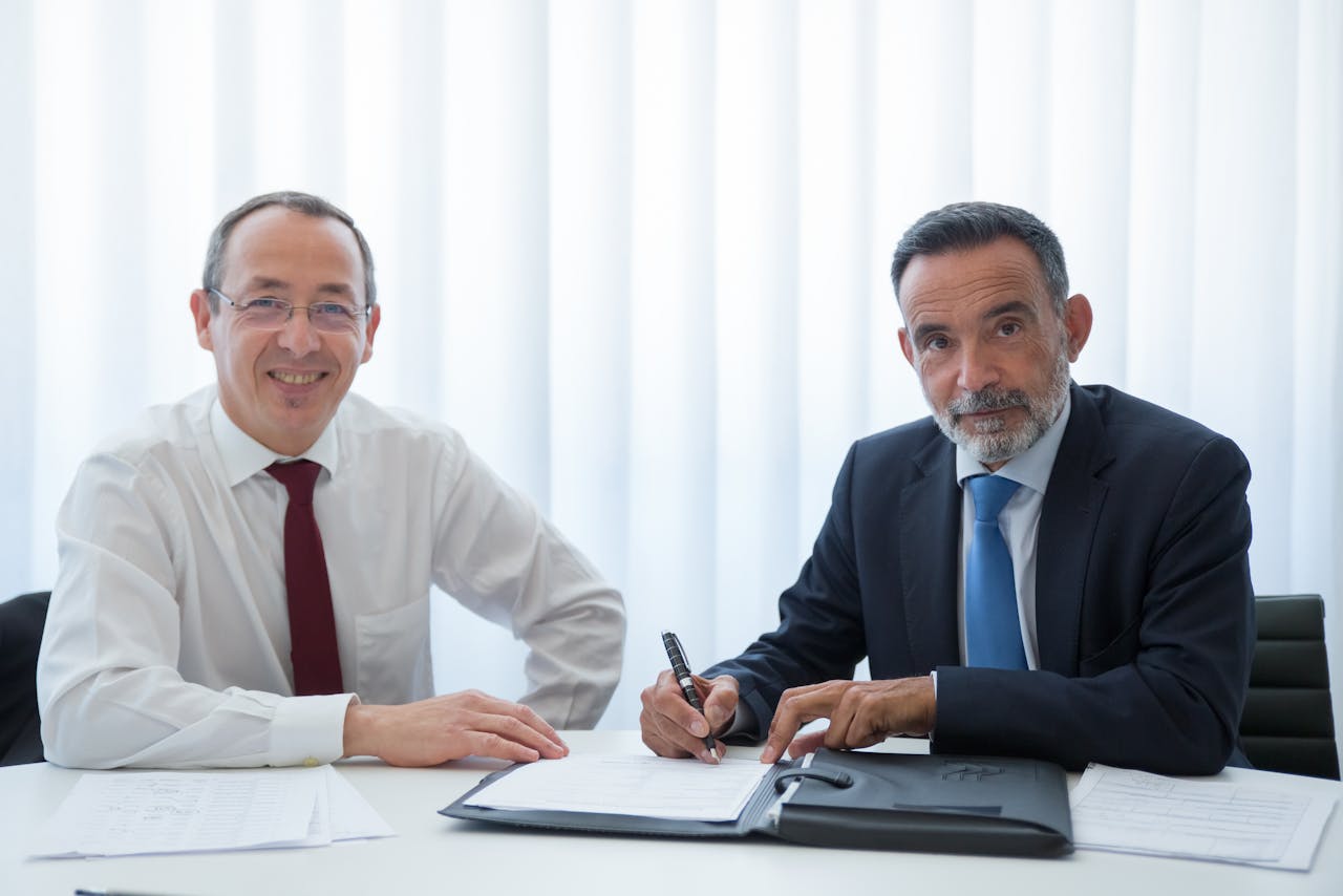 Two businessmen discussing and signing documents at a bright office table.